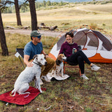 A couple with two dogs on Layzee Dog My Place dog beds sitting in front of a tent in a natural setting