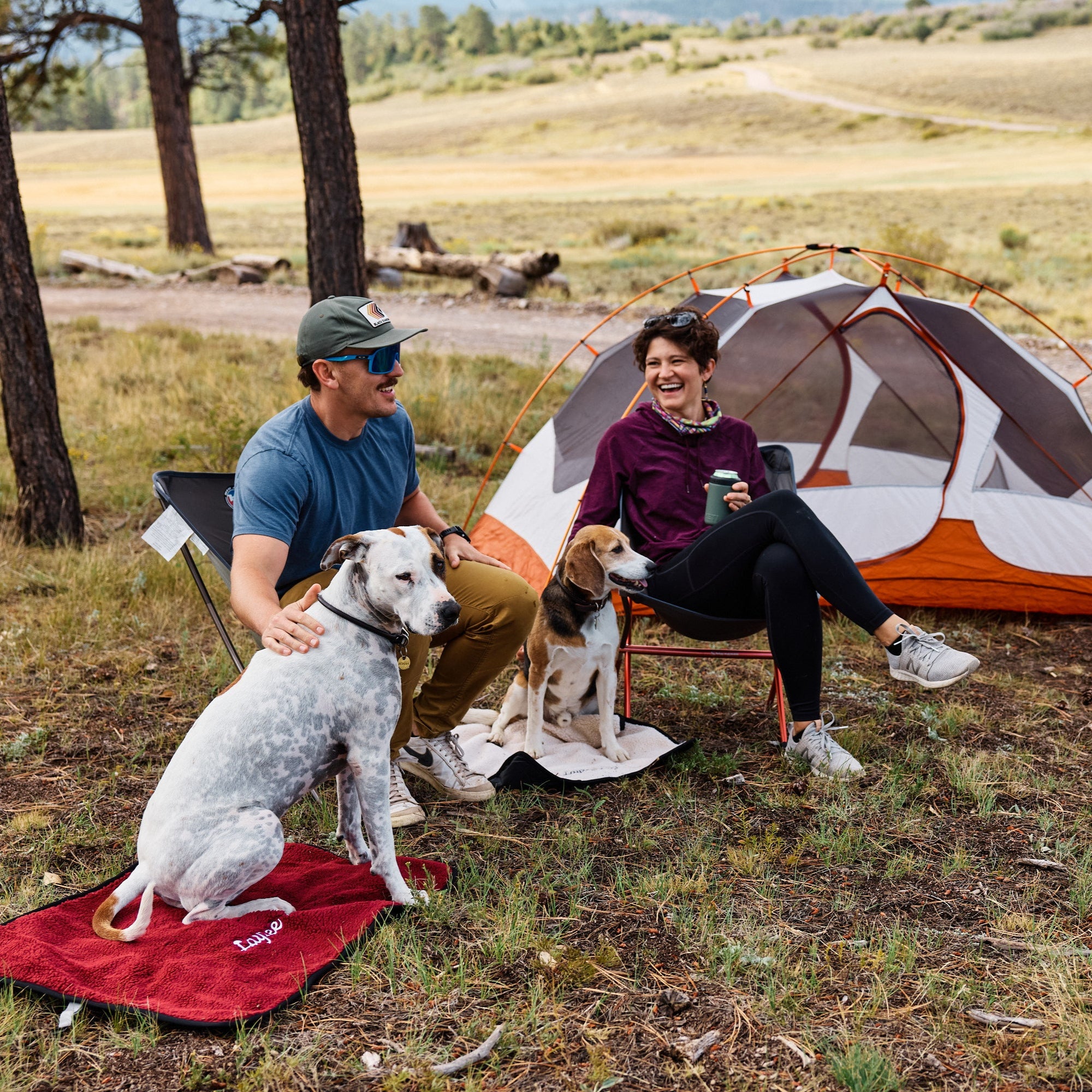 A couple with two dogs on Layzee Dog My Place dog beds sitting in front of a tent in a natural setting
