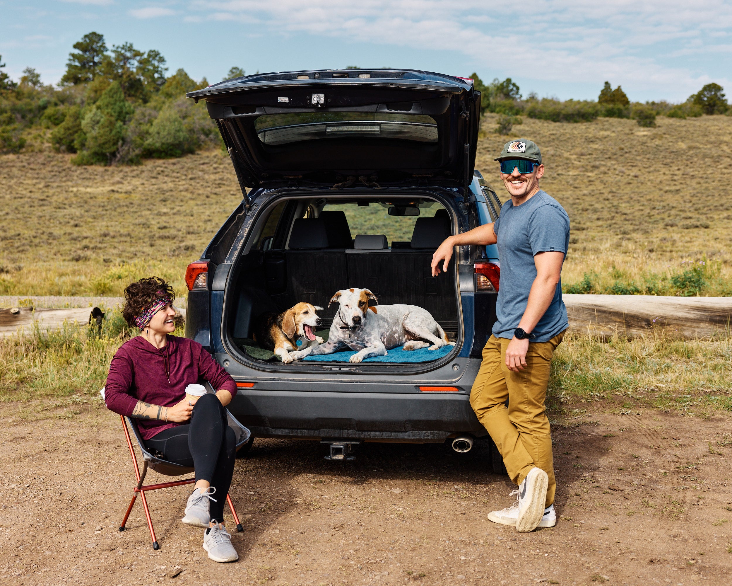 Two people with two dogs lying on My Place portable dog beds in the back of an open car trunk outdoors