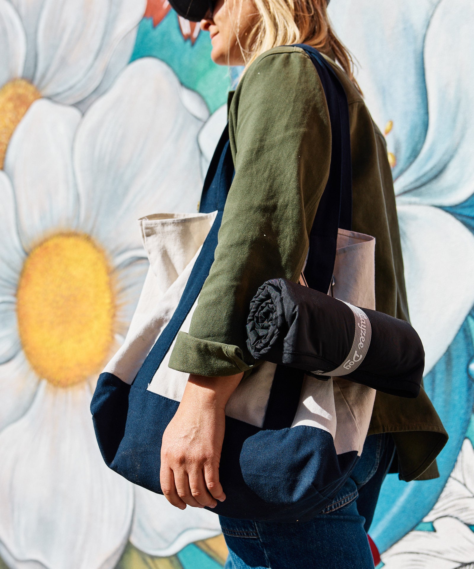 A person with a My Place ultralight, portable dog bed rolled up and clipped to a tote bag with a colorful, floral mural in the background